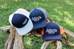 Three hats with 'The Ingram Distillery' logo on a wooden log against a grassy background